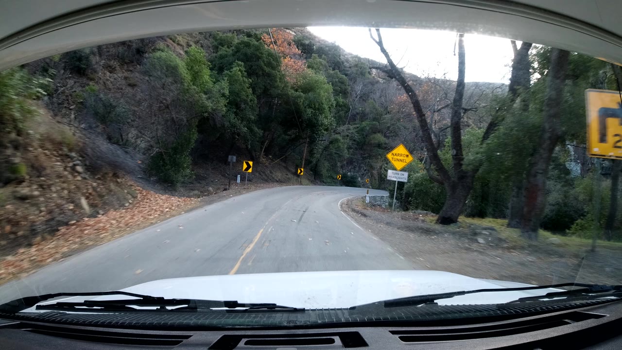 In Ojai, California, the vehicle approaches a right bend on a two-lane road bordered by trees, dry leaves, a hillside shoulder, yellow warning signs, and no visible people.