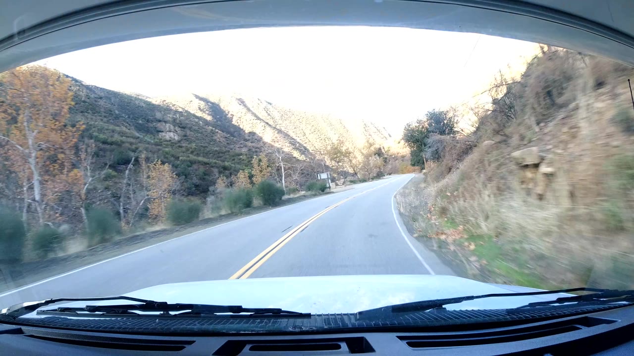 A dashboard view in Ojai, California, shows a sunlit two-lane road climbing beside dry slopes, shrubs, a yellow warning sign near the windshield edge, and no visible people.