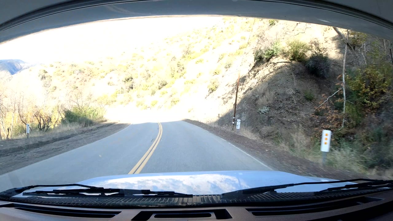 In Ojai, California, the windshield frames a left-curving two-lane road beside a bright exposed hillside, roadside posts, sparse brush, and no visible people.