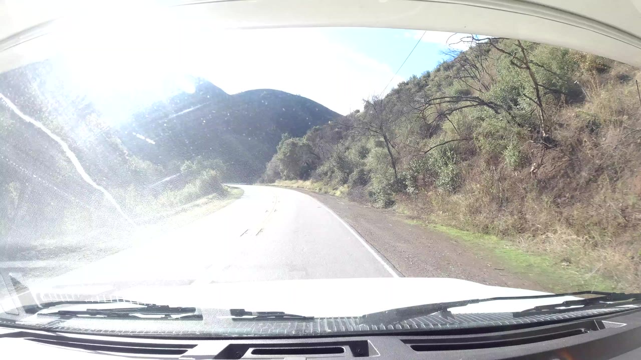 Opening frame of an empty Ojai, California road curving through dry hills, with windshield glare, car hood, and no visible people.