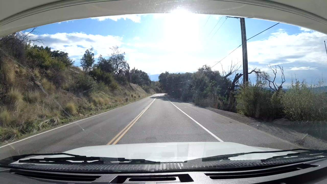 Windshield view in Ojai, California, looking down a two-lane road bordered by dry hillside brush, trees, utility poles, and bright sky, with no visible people.