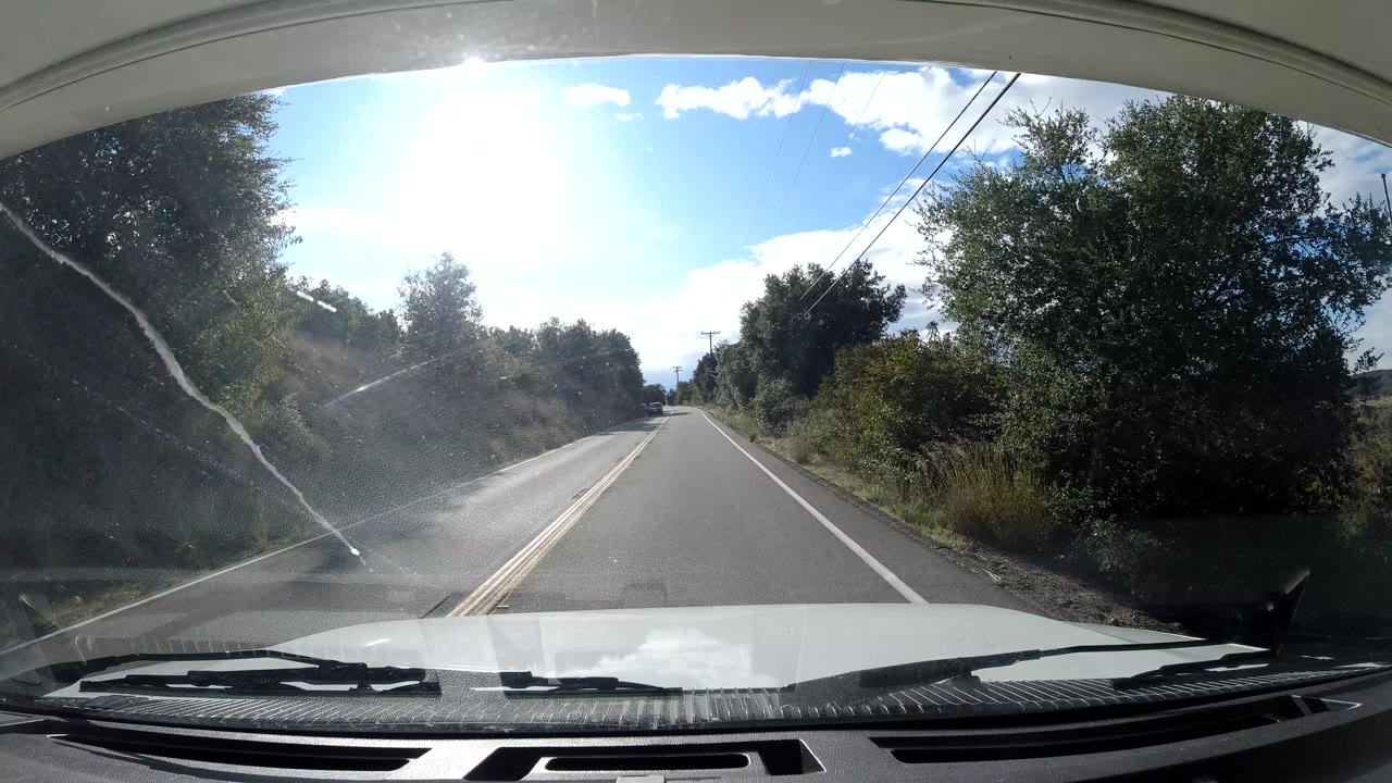 Ojai, California windshield view along a straighter two-lane road with trees and brush on both sides, overhead utility lines, bright sun, and no visible people.