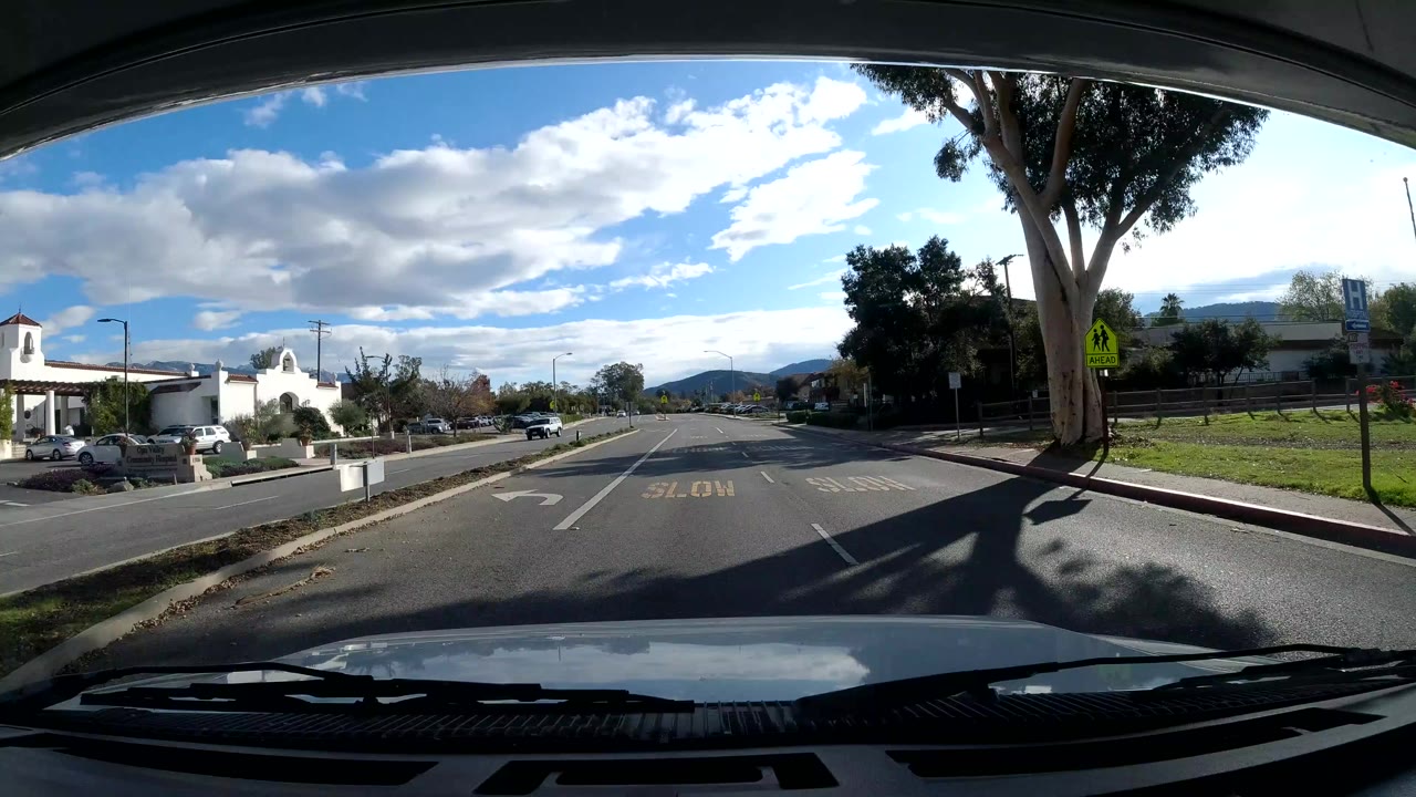 Forward windshield view on an Ojai, California road with lane markings, trees, a white building to the left, distant hills, scattered clouds, and no visible people.