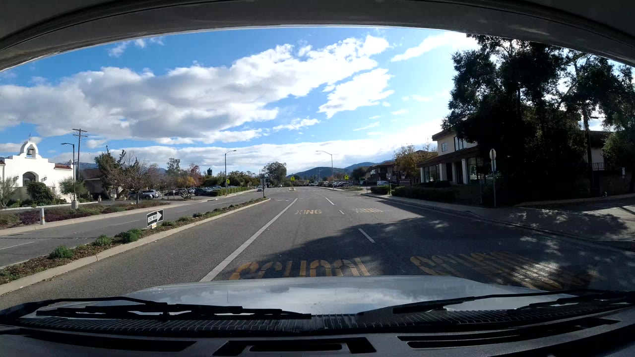 Vehicle POV down a road with a center lane, trees, buildings, and mountains ahead.