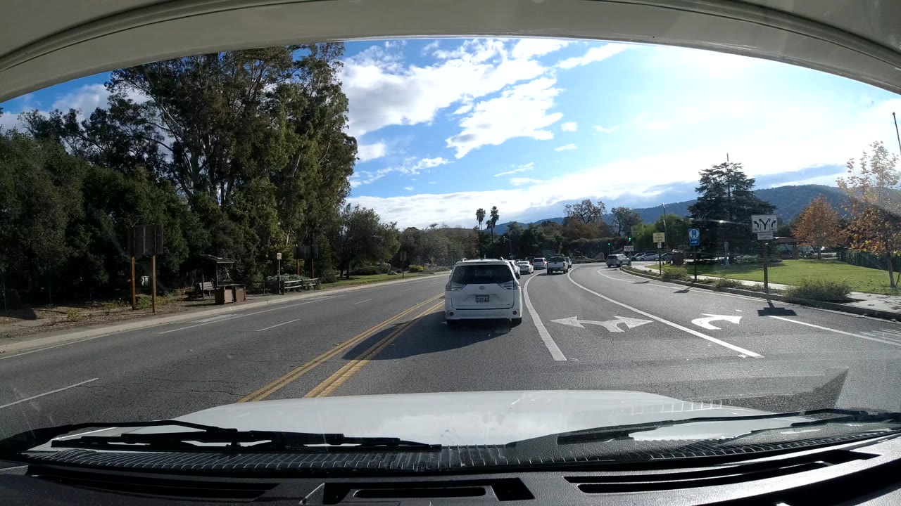 Forward view through a windshield in Ojai, California showing a white vehicle ahead near road arrows, trees along the roadside, distant mountains, bright sky, and no visible people.