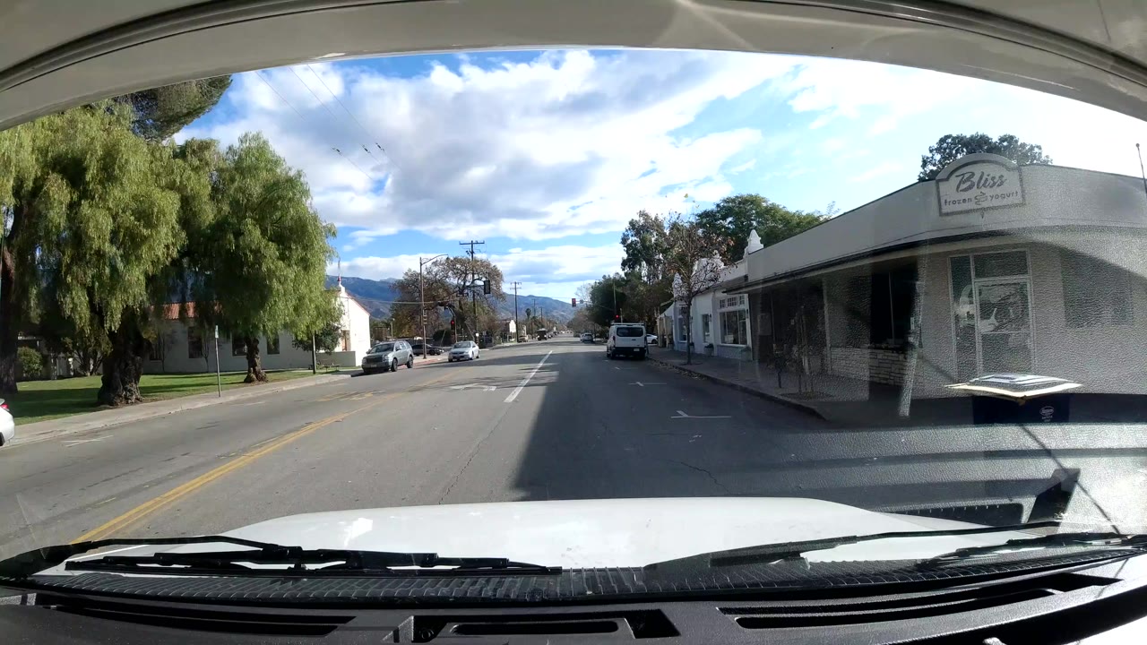 Vehicle-mounted view in Ojai, California looking down a neighborhood street past a storefront, parked cars, leafy trees, bright clouds, and distant hills, with no clearly visible people.