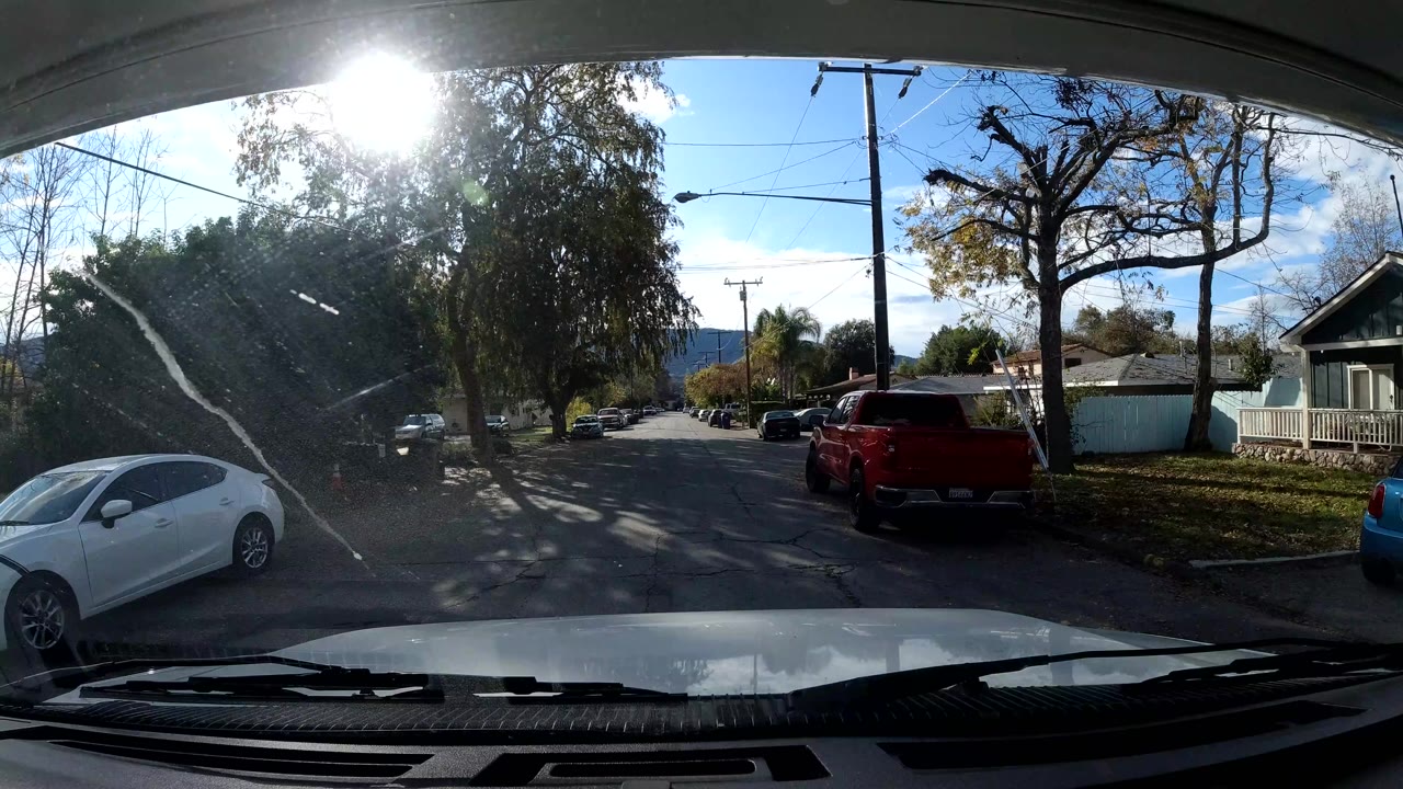 Vehicle view in Ojai, California angled toward a side street where a white car sits near the curb, with homes, trees, utility lines, parked cars, sun glare, and no clearly visible people.