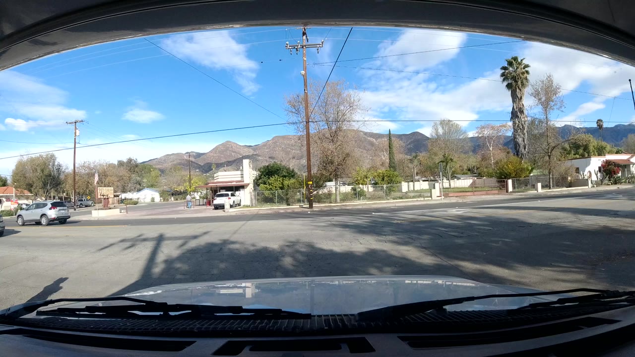 At 0:01, the vehicle view remains on a broad Ojai street with low roadside buildings, parked cars, trees, power lines, and mountains behind the neighborhood, with no clearly visible people.
