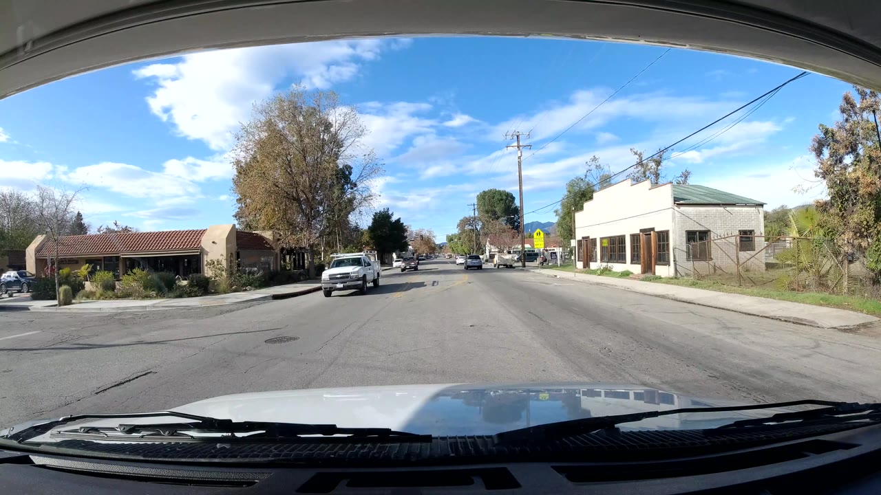 At 0:02, the vehicle faces down an Ojai street with a white truck ahead, small roadside buildings, a pedestrian crossing sign, utility lines, trees, and bright blue sky, with no clearly visible people.