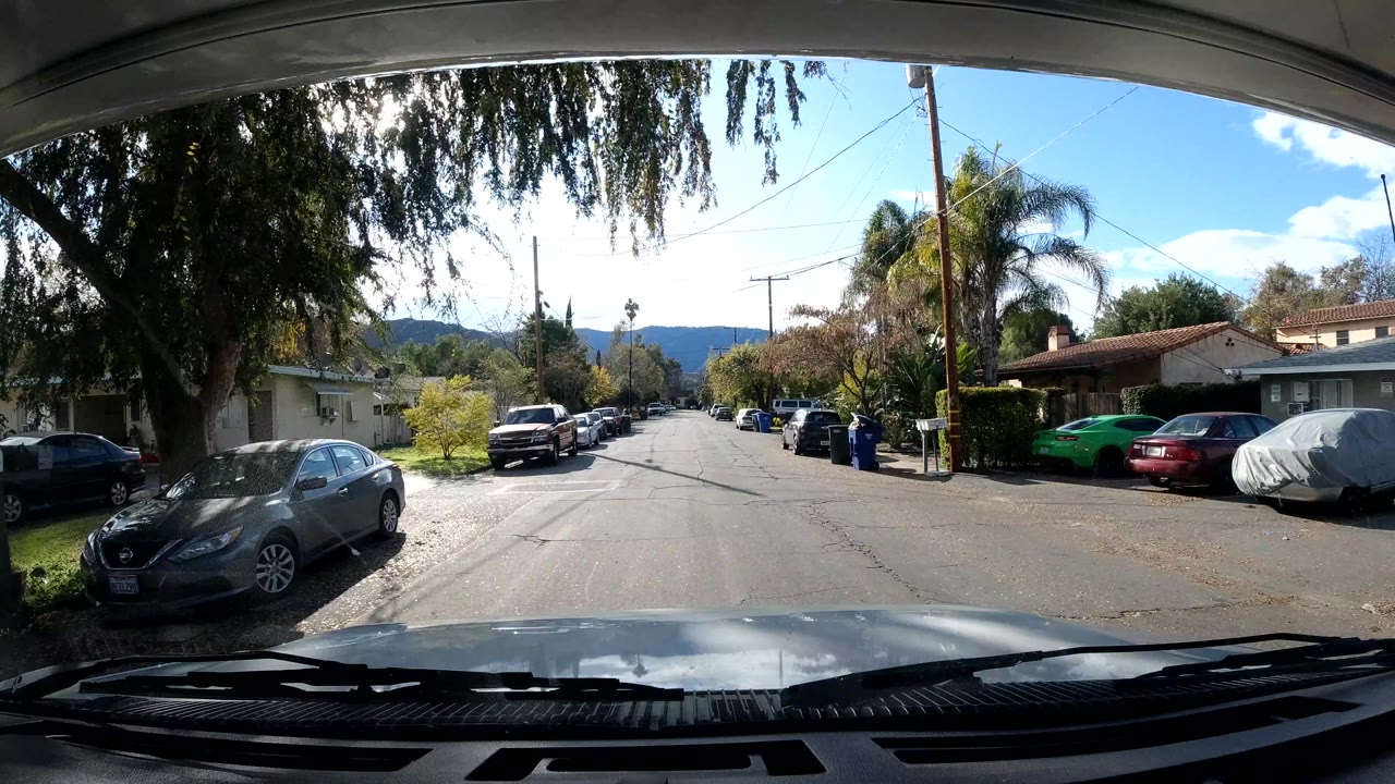Residential street seen from a vehicle, with parked cars, trees, homes, and mountains beyond.