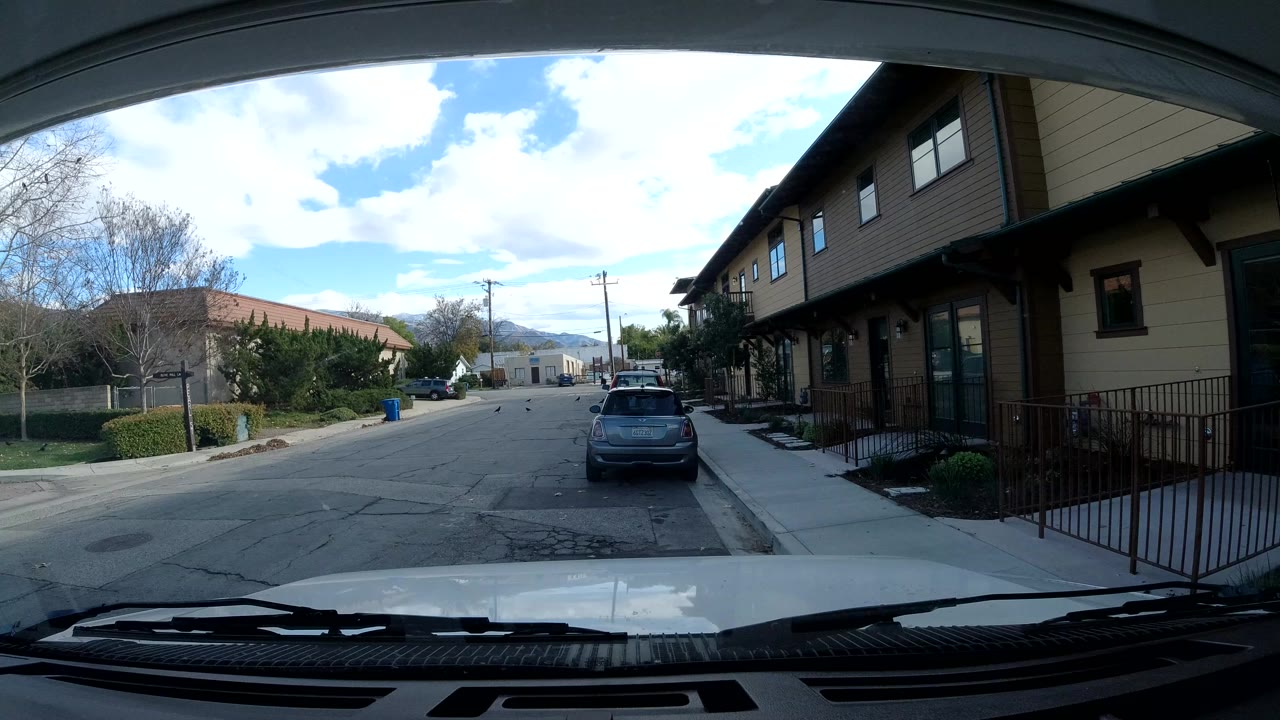 Opening frame of an Ojai, California neighborhood street seen over a vehicle hood, with a parked hatchback ahead, tan buildings on the right, trees on the left, and no visible people.