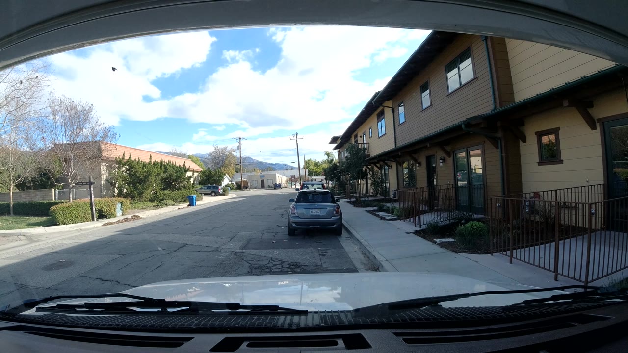 Mid-clip Ojai neighborhood view showing the parked hatchback near center, a sidewalk and railing along the right building, open street pavement, and no visible people.