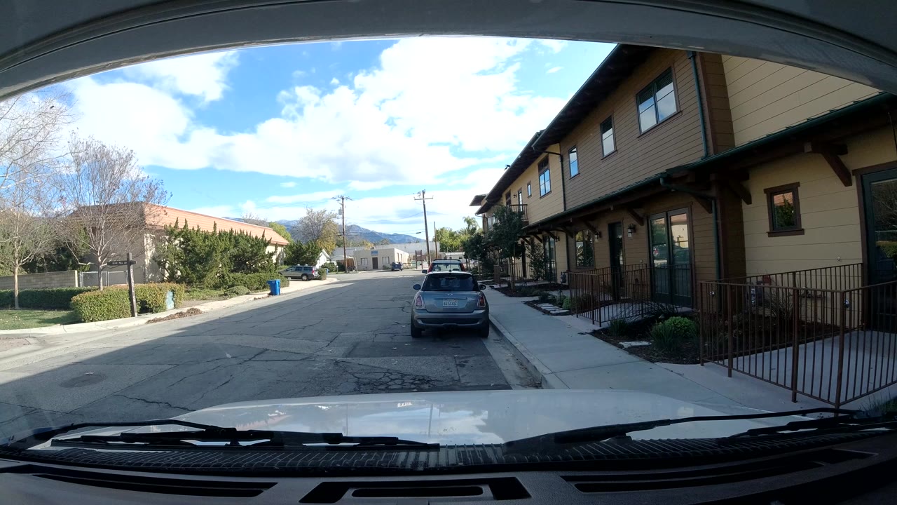 Dashboard view in Ojai, California looking down a quiet street with tan buildings at right, a parked car ahead, trees and distant hills under bright sky, with no visible people.