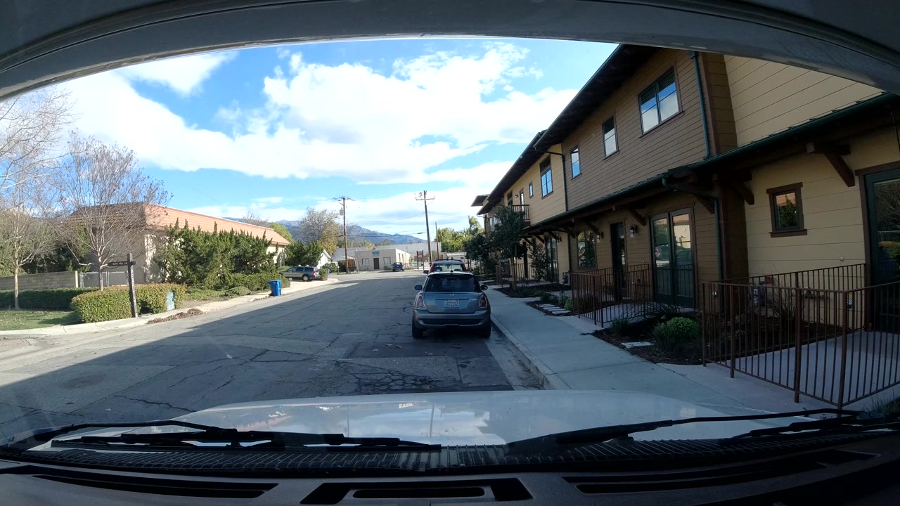 Opening frame of an Ojai, California street seen through a windshield, with a parked hatchback ahead, beige building on the right, trees on the left, distant hills, and no visible people.