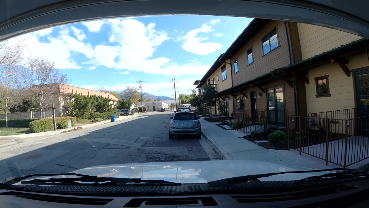 Ojai, California street view from inside a vehicle, with the dashboard at the bottom, a parked car centered ahead, tan siding and railings on the right, trees to the left, and no visible people.