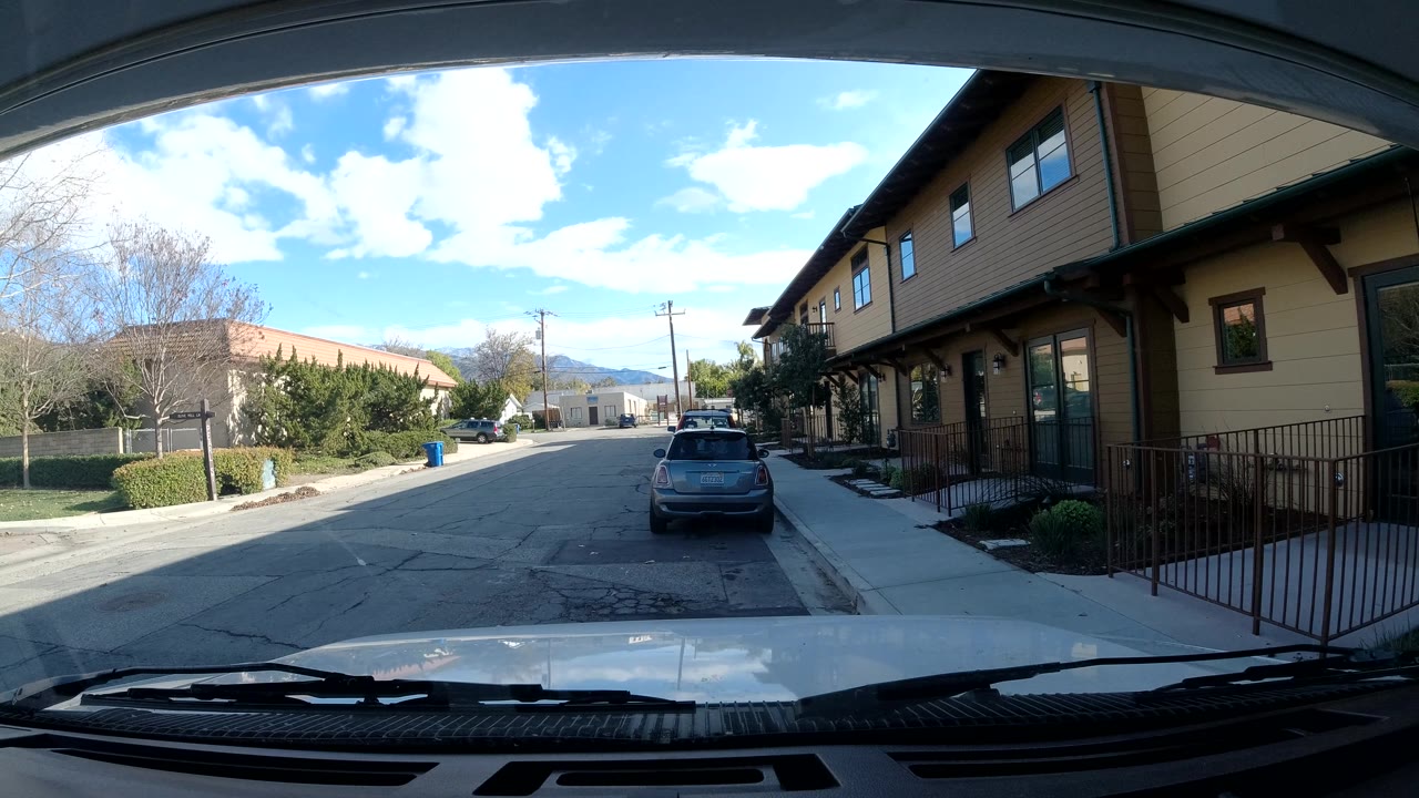 Dash-cam street scene with sidewalk railing and parked hatchback.