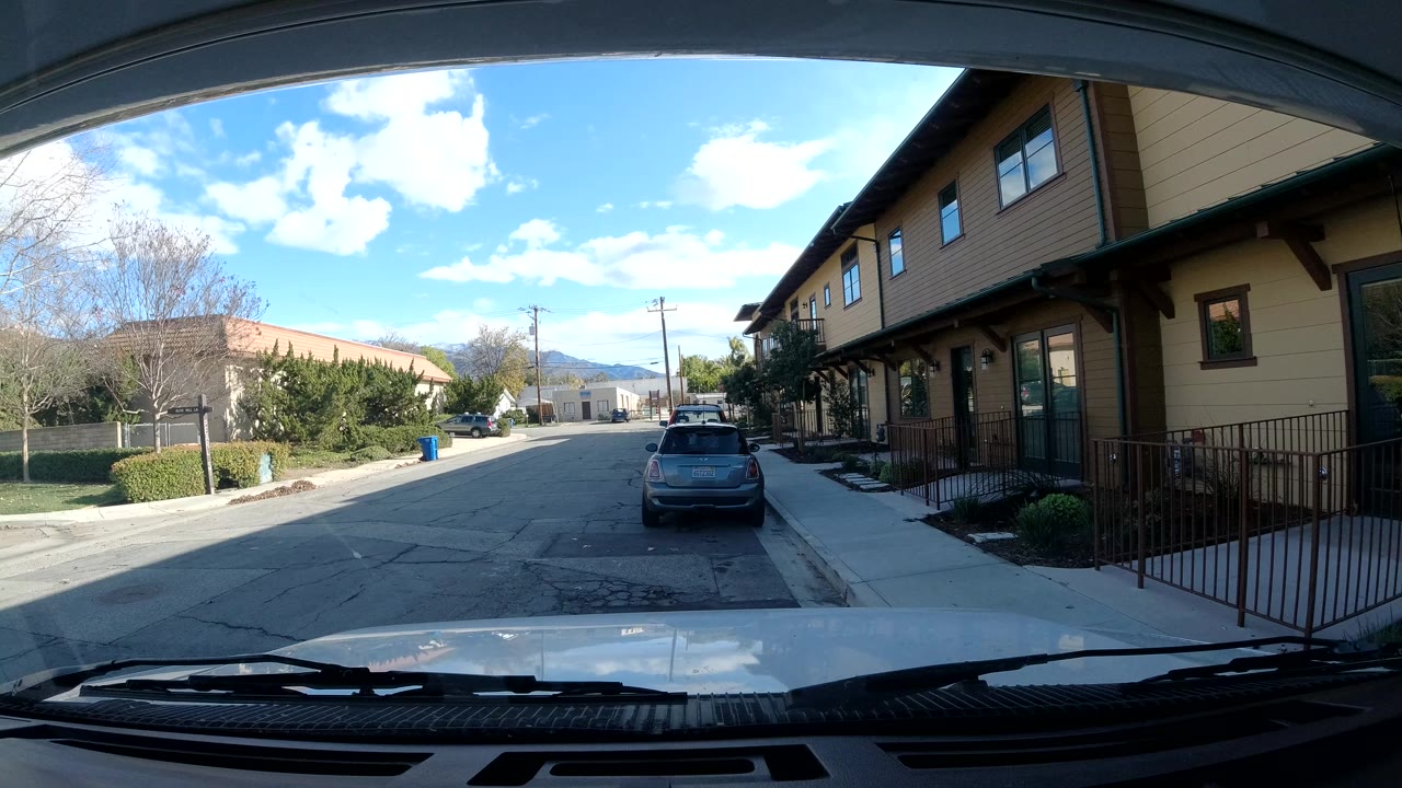 Opening frame of an Ojai, California street viewed from behind a windshield, with a parked hatchback ahead, tan building exterior on the right, trees on the left, blue sky, and no visible people.