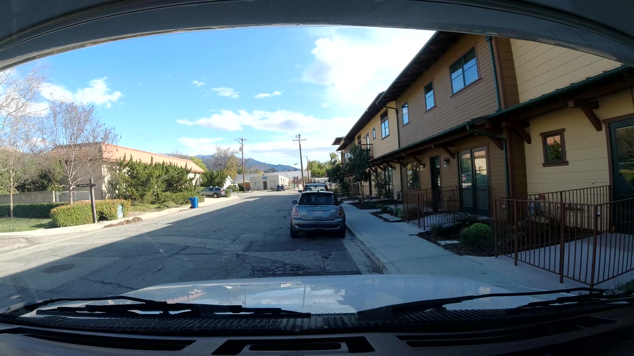 Parked-car windshield view with street, gray car, sidewalk railing, and tan building facade.