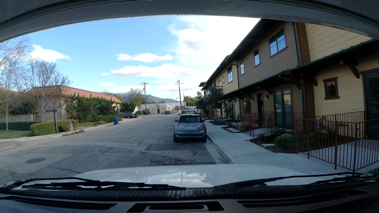 Second view of the Ojai street keeps the parked car centered, with beige residential buildings on the right, a sidewalk and railings beside them, and no visible people.