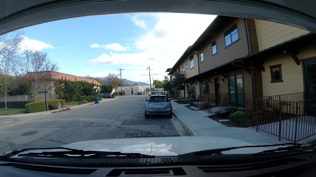 Vehicle windshield view of a residential block with a gray car parked ahead.