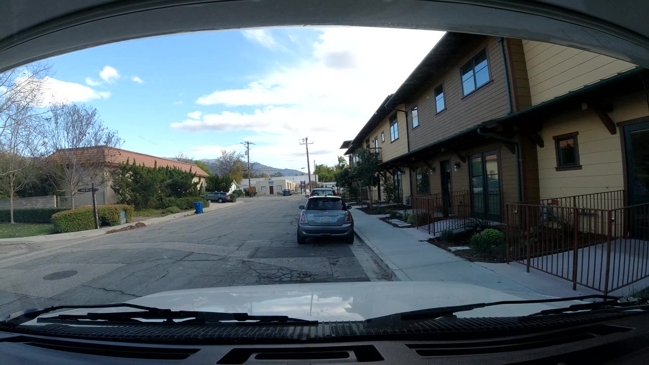 Ojai, California residential street viewed over a vehicle dashboard, with a dark parked car ahead, tan homes on the right, trees on the left, and no visible people.