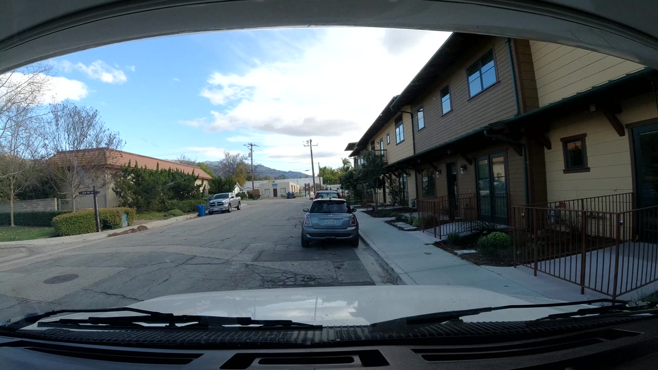 Opening Ojai street view from behind a vehicle windshield, with hood and wipers at the bottom, parked cars ahead, tan building facades on the right, distant hills, and no visible people.
