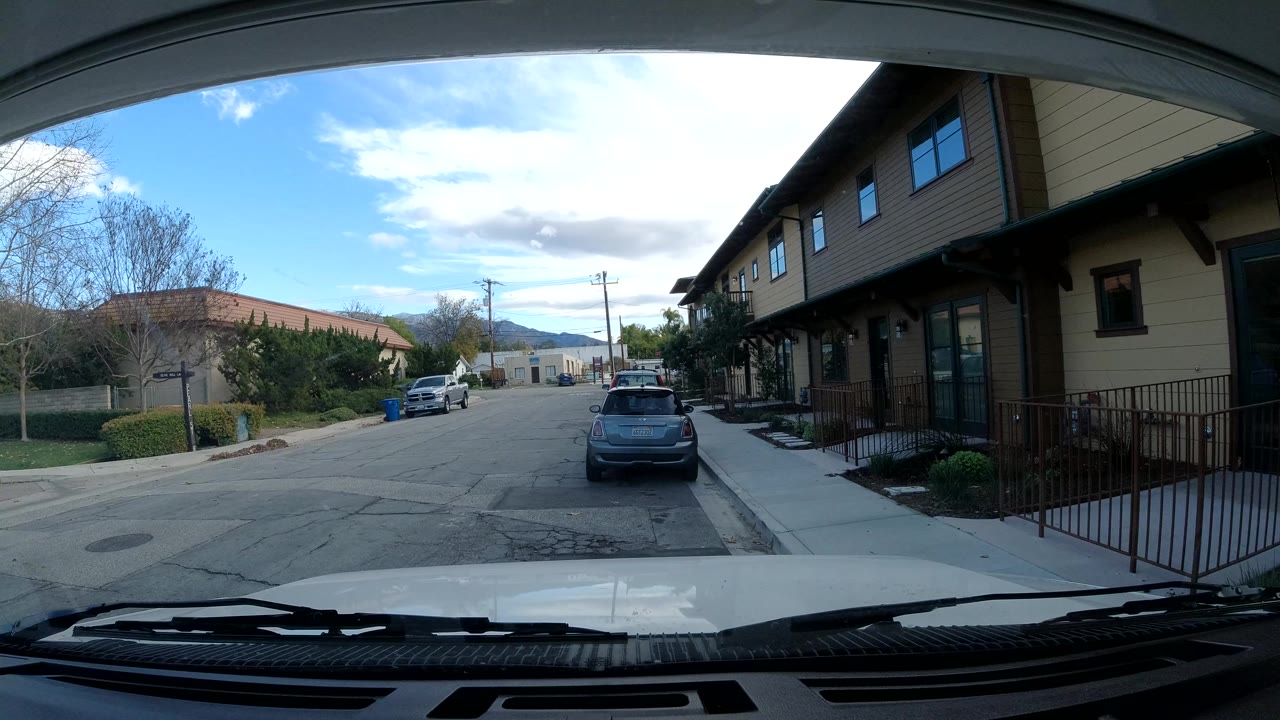 Opening frame of an Ojai, California side road viewed through a vehicle windshield, with parked cars, condo buildings, trees, utility lines, distant hills, and no visible people.