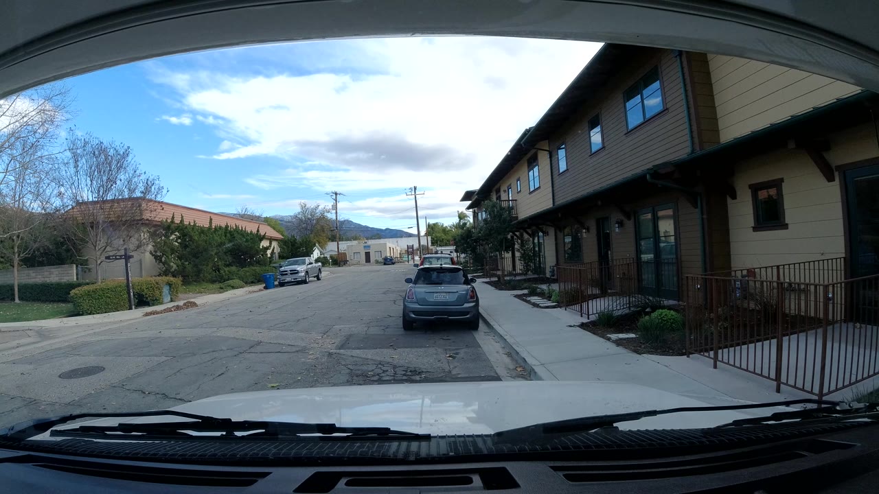 Forward windshield view in Ojai, California, showing a stopped vehicle perspective on a street with parked cars, a tan building on the right, trees on the left, distant hills, and no visible people.