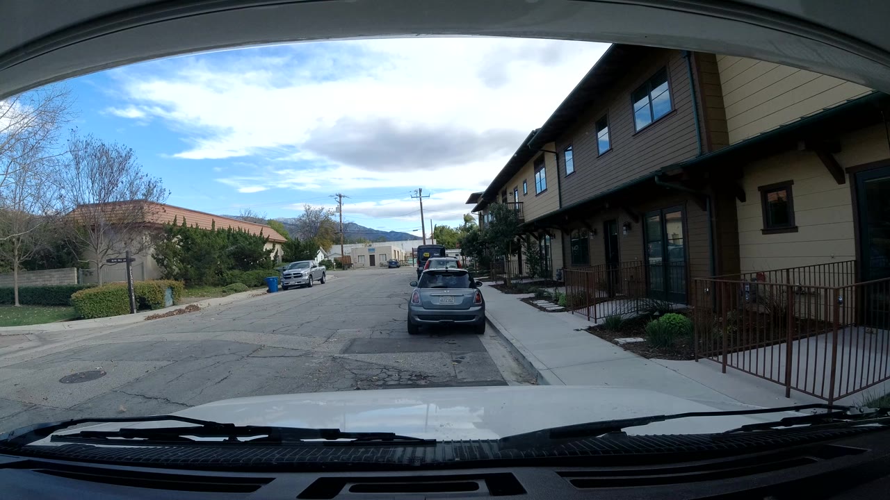Opening view of an Ojai, California residential street from behind a windshield, with a parked hatchback ahead, tan homes at right, trees at left, blue sky, and no visible people.