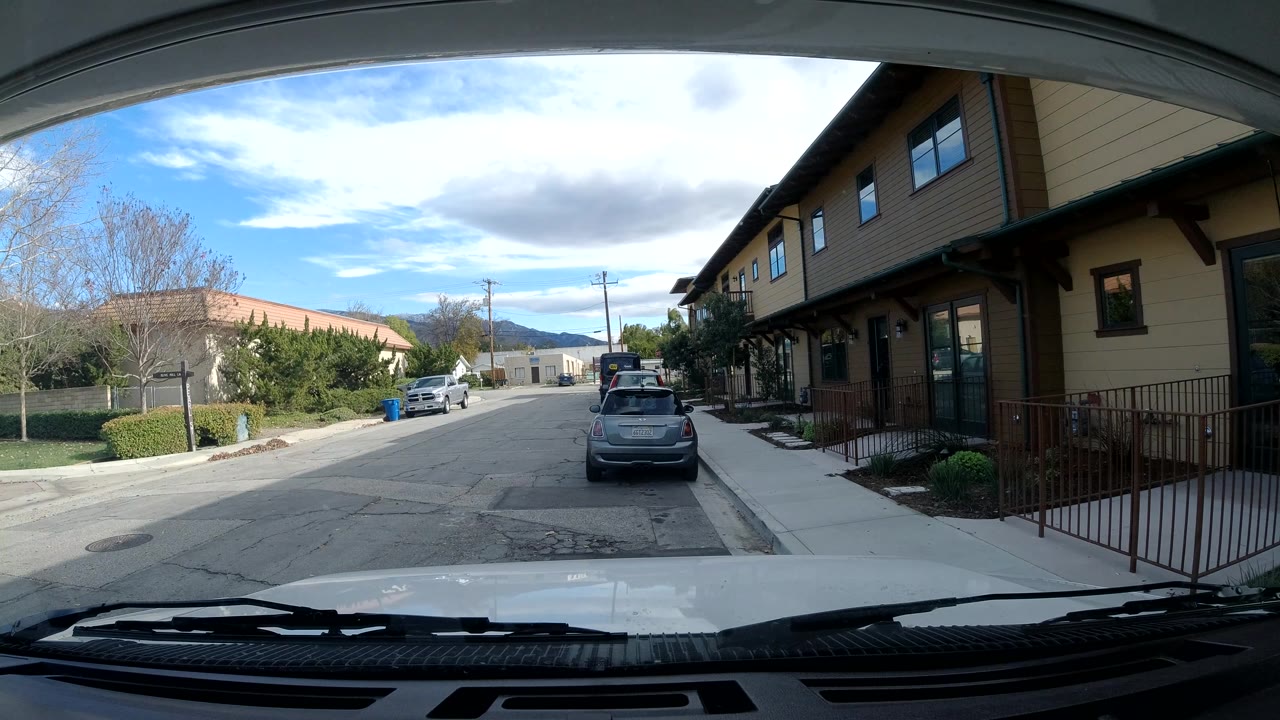 The third frame keeps the Ojai residential lane centered, with the hatchback parked ahead, tan buildings at right, landscaped greenery at left, distant hills, and no visible people.