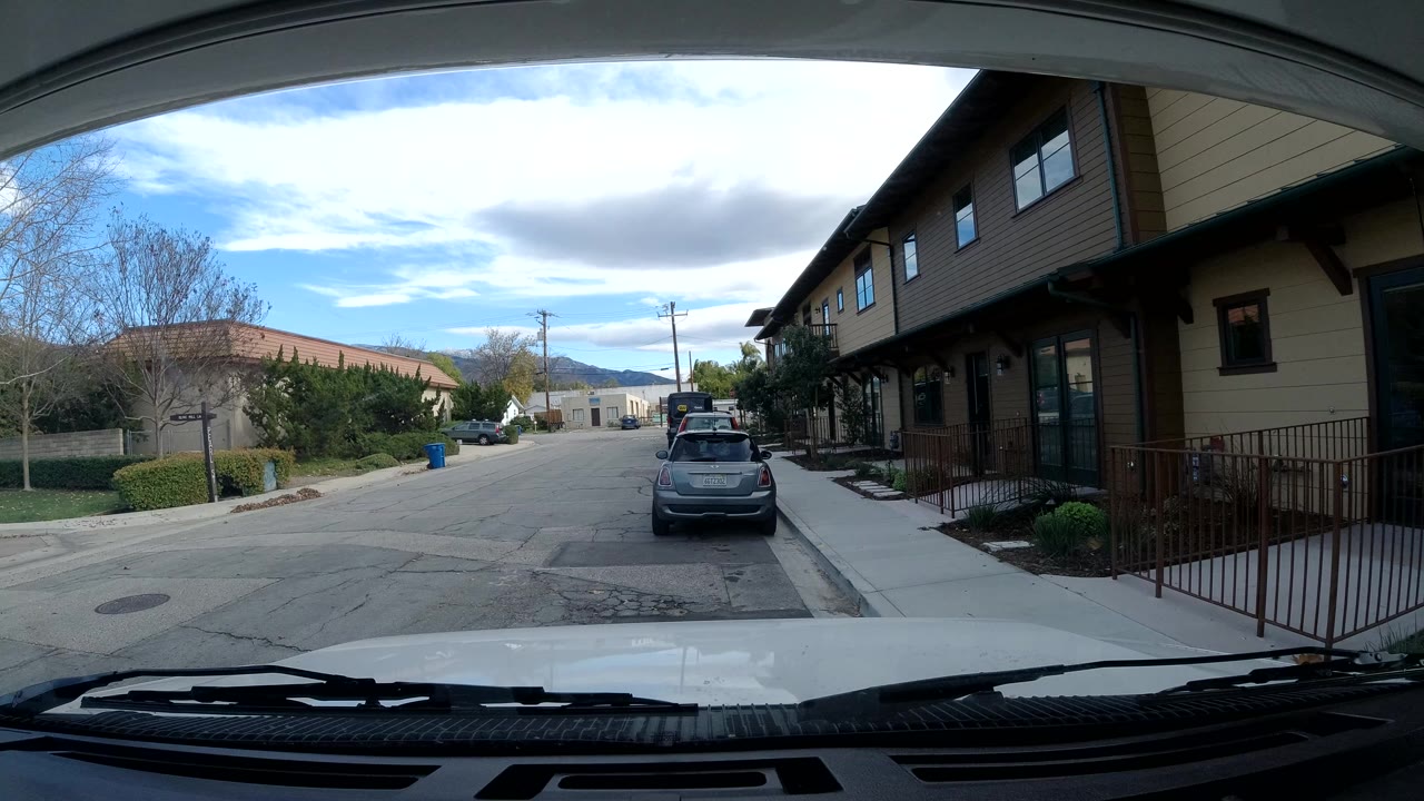 Opening Ojai windshield view with dashboard and wipers in the foreground, a parked gray car ahead, tan buildings along the right sidewalk, trees at left, distant hills, and no visible people.