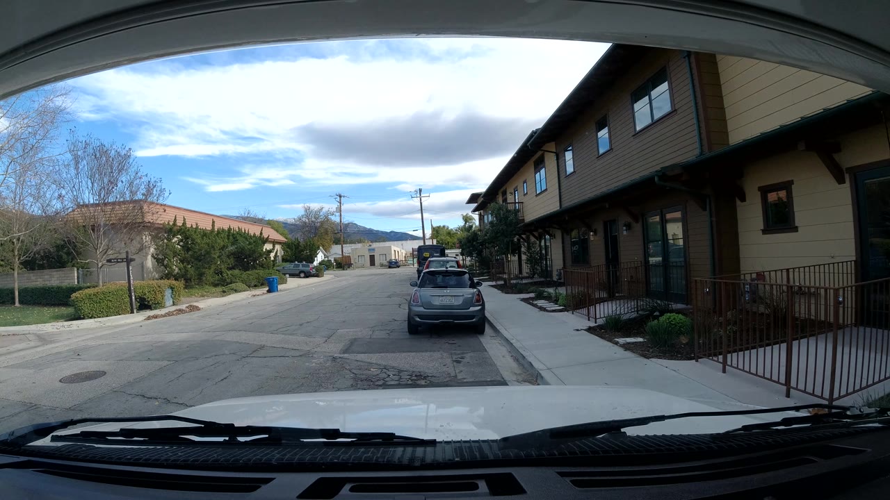 At 0:02, the Ojai street remains framed by the car interior, with the parked gray car slightly right of center, leafless trees and hedges at left, tan buildings at right, and no visible people.