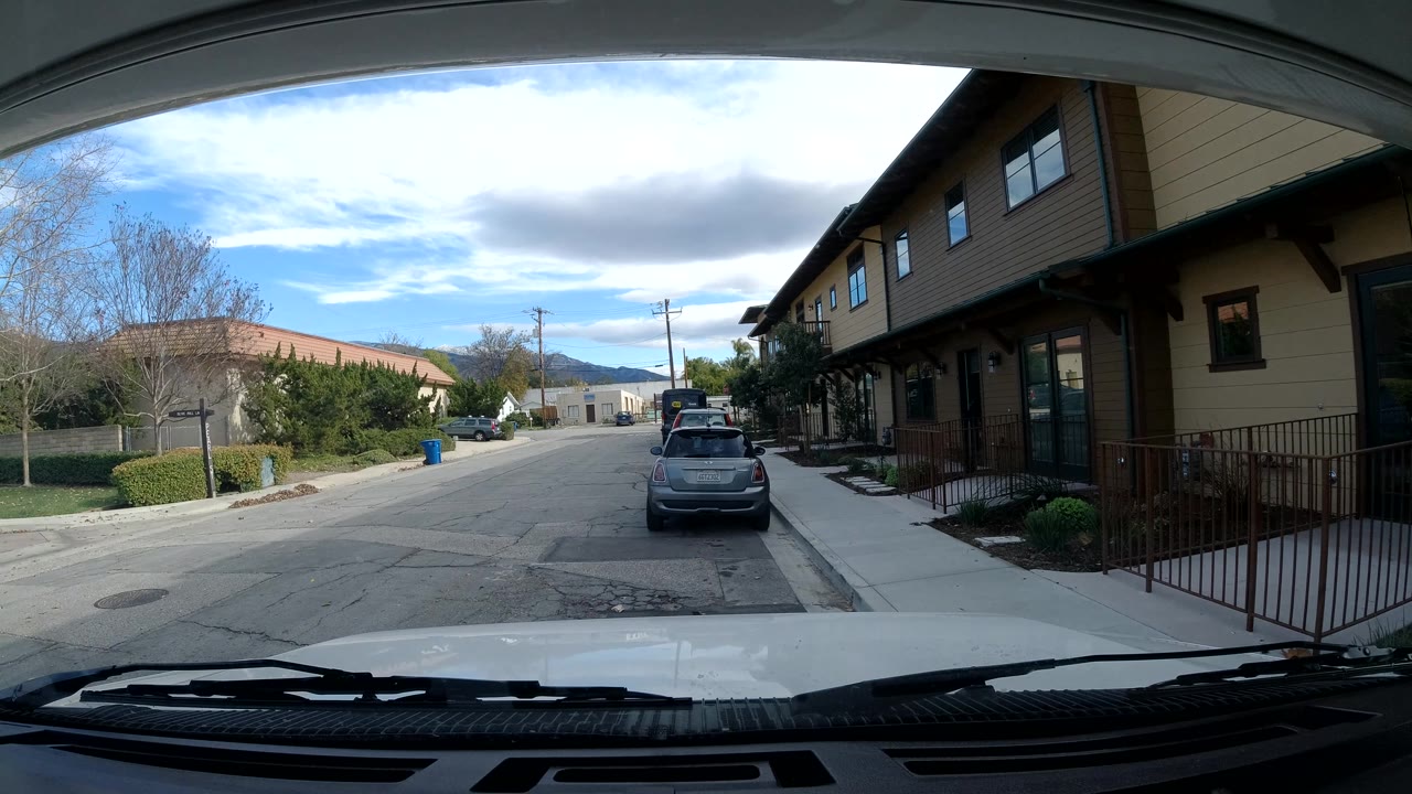 Interior car POV of a quiet street with sidewalk, railing, parked car, and cloudy sky.