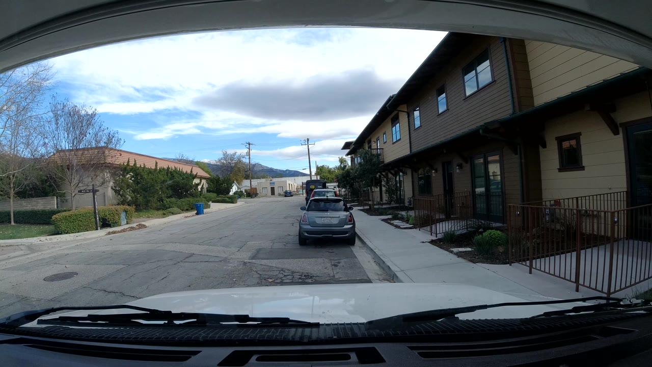 Opening frame from Ojai, California, showing a windshield-framed residential street, a gray parked car, tan buildings, trees, utility poles, distant hills, and no visible people.
