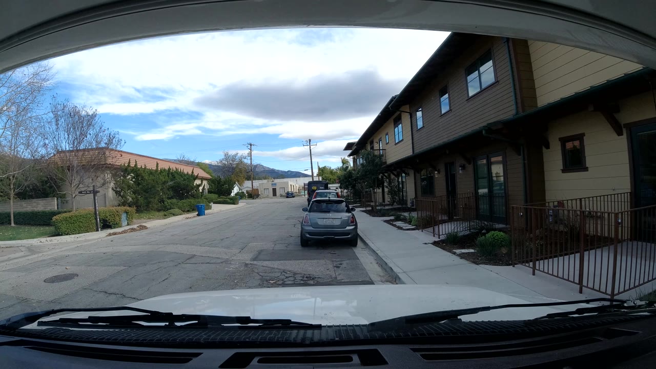 Second frame from Ojai, California, with the car hood low in frame, a parked gray car centered ahead, residential buildings to the right, trees to the left, and no visible people.