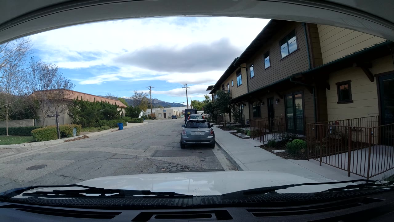 Third frame from Ojai, California, showing the parked car slightly right of center, tan residential structures along the sidewalk, bare trees, overhead clouds, and no visible people.