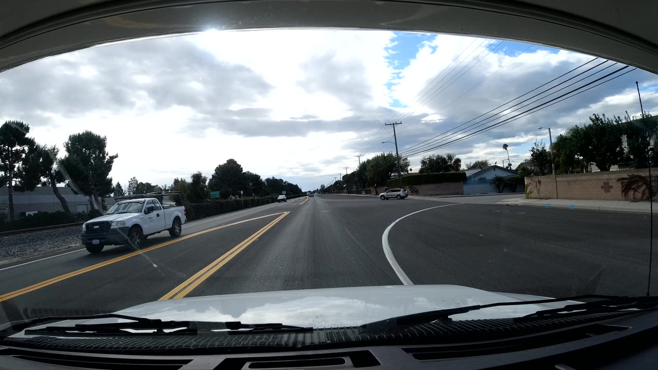 Forward windshield view on a Malibu road with a white pickup approaching at left, roadside trees, power lines, a low wall, scattered clouds, and no visible people.