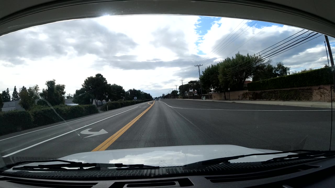 Vehicle-mounted view along a Malibu road with yellow center lines, a right-turn lane marking, trees behind roadside walls, overhead wires, bright clouds, and no visible people.