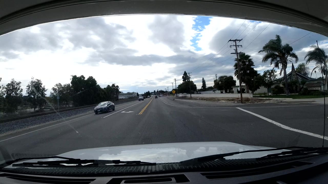 Malibu street intersection seen through the windshield with several cars, palm trees, power poles, road markings, low buildings, bright clouds, and no visible people.