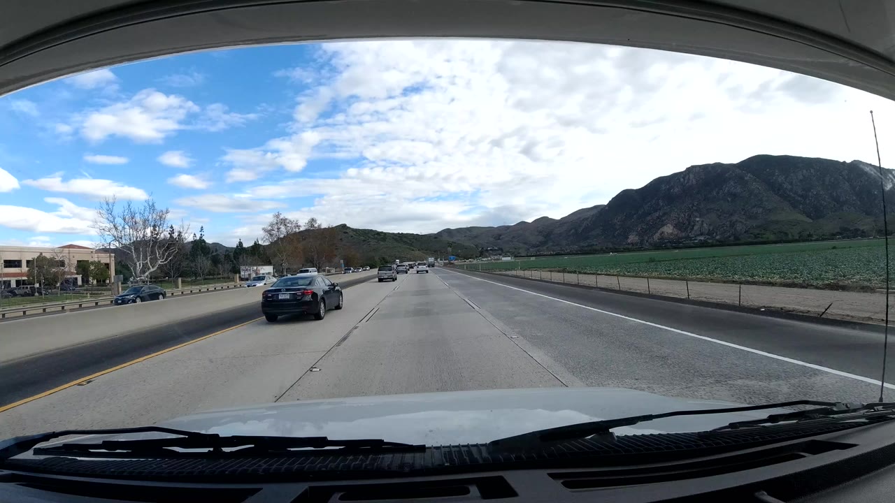 Dashcam view in Malibu, California, showing a multi-lane highway, dashboard foreground, cars ahead, green hills, blue sky, and no visible people.