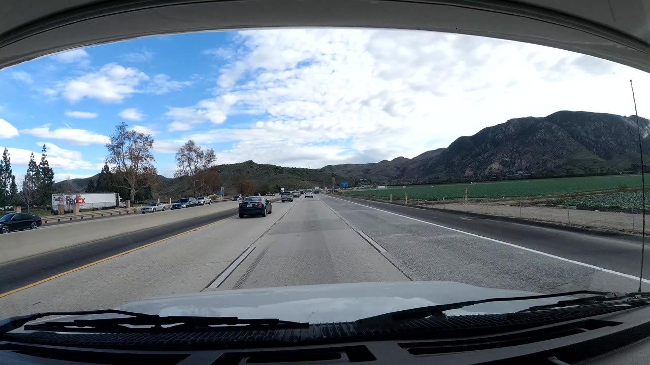 Forward road view in Malibu, California, with a dashboard edge, highway lanes, passenger cars, a branded truck in the distance, hillside fields, and no visible people.