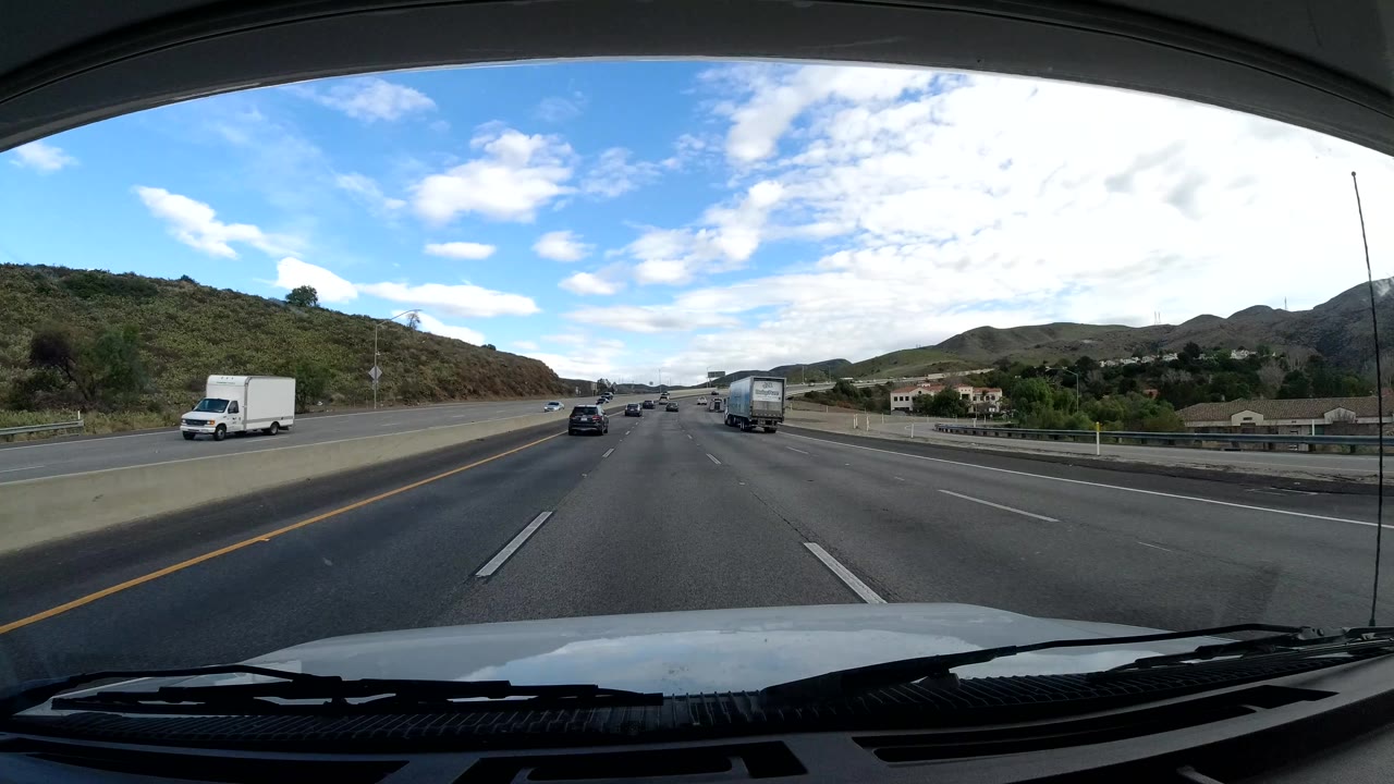 Dashcam frame in Malibu, California, showing an open multi-lane road, cars and box trucks, rolling hills, roadside buildings, blue sky, and no visible people.