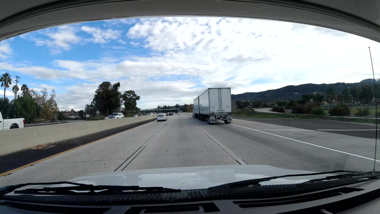 Forward windshield view of Malibu freeway lanes with a white box truck ahead, cars in adjacent lanes, roadside trees, distant hills, broken clouds, and no visible people.