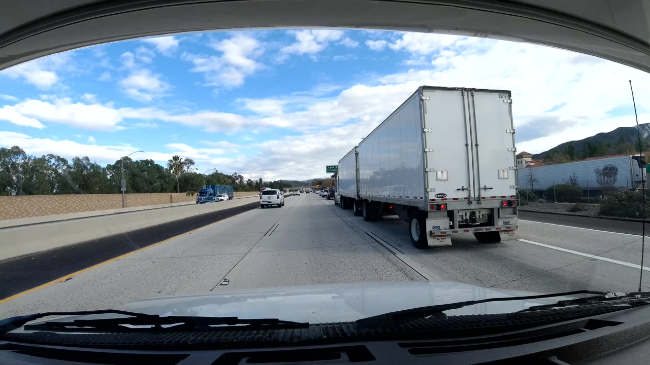 Malibu freeway view with a large white box truck close on the right, cars ahead, a concrete barrier, hillside greenery, blue sky, and no visible people.
