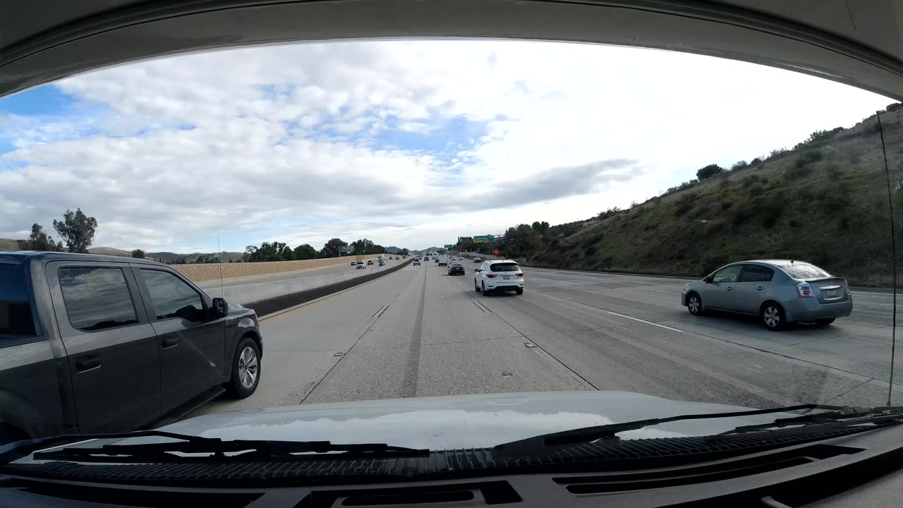 Forward-facing Malibu freeway view from a vehicle, with several cars ahead, a grassy hillside to the right, partly cloudy daylight, and no visible people.