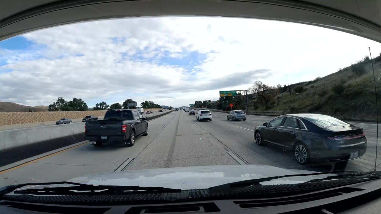 Malibu road-travel frame with a black pickup and dark sedan near the camera, a concrete divider, hillside brush, overhead road sign, and no visible people.