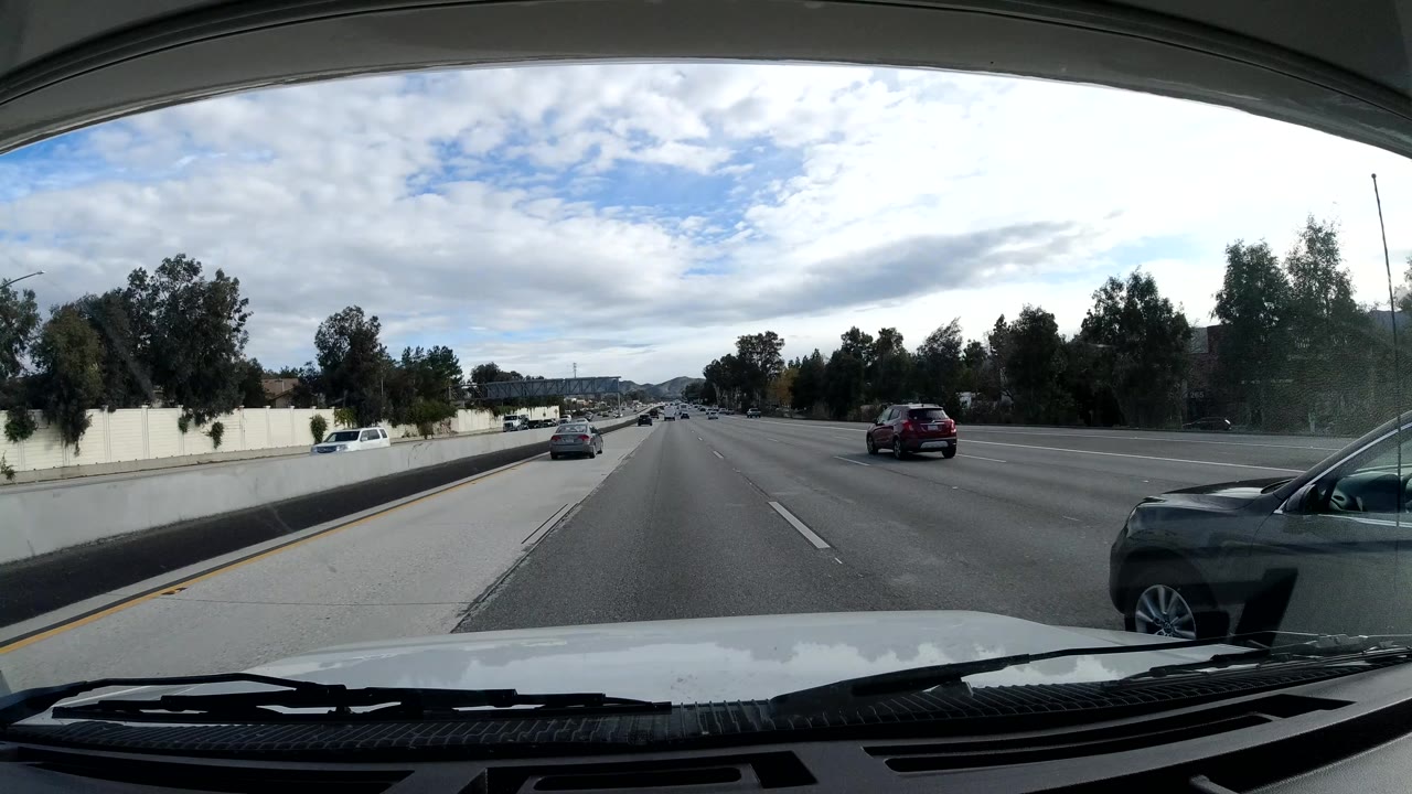 Forward Malibu freeway view with traffic spread across wide lanes, a concrete median wall, roadside trees, distant hills, bright cloud cover, and no visible people.