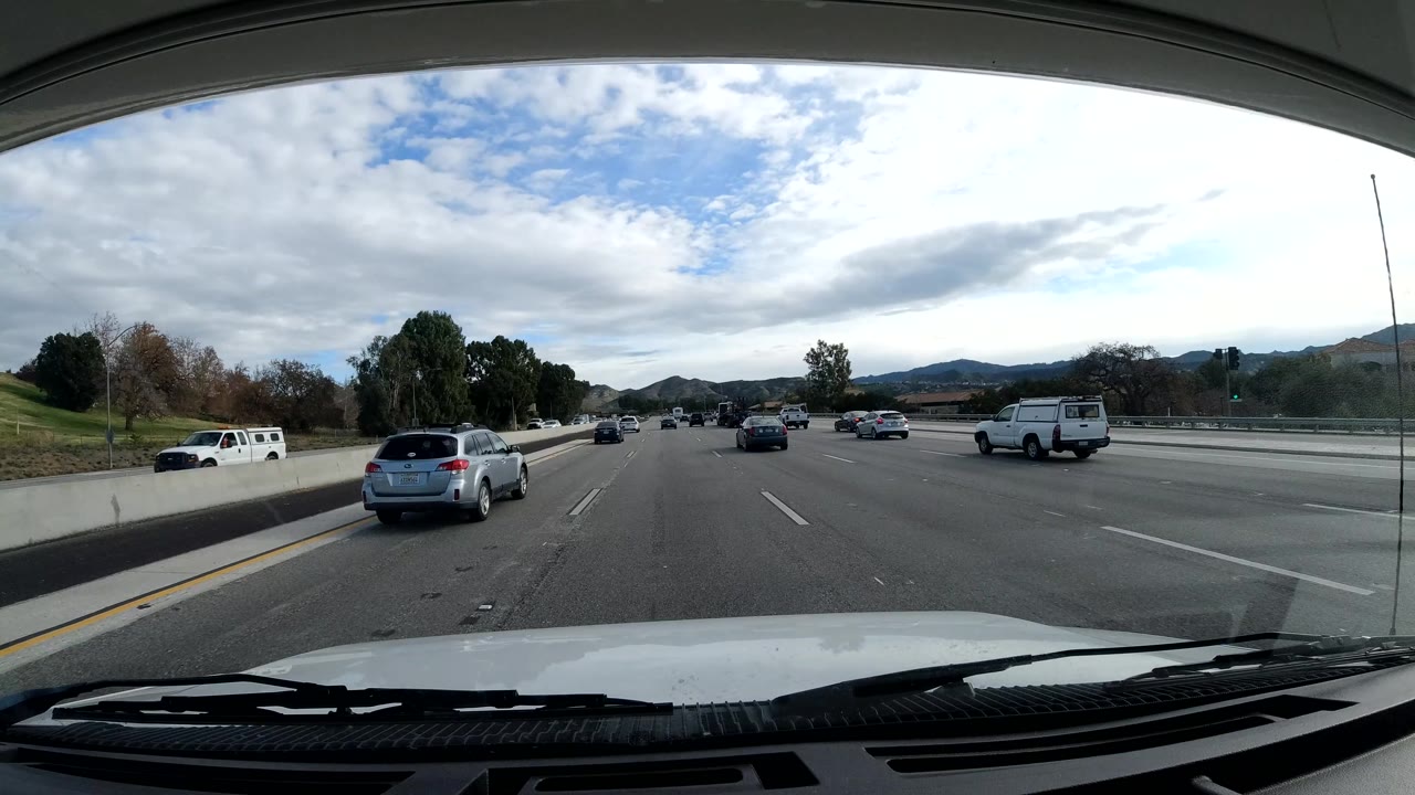 Freeway traffic beside trees and hills with vehicle hood at the bottom of frame.