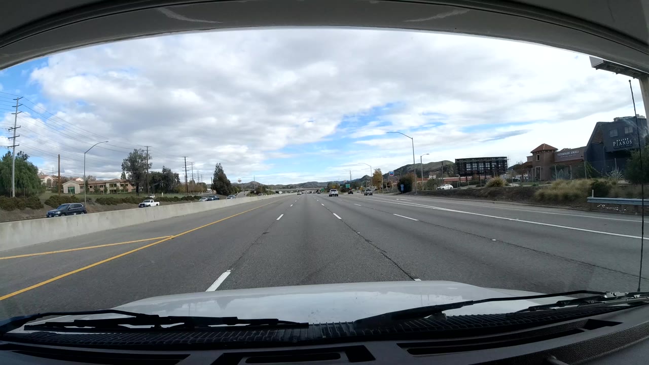 Second forward road view in Malibu, California with the hood at the bottom of frame, low commercial or residential buildings to the right, light traffic ahead, hills, and no visible people.