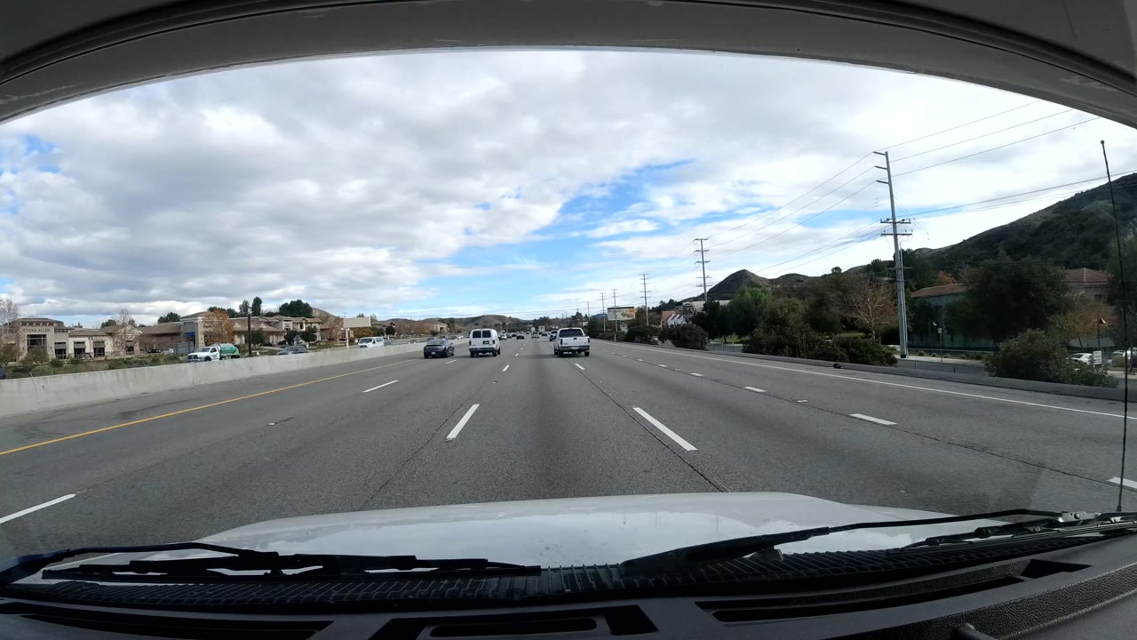 Forward-facing Malibu, California road frame with several cars ahead, a hillside on the right, roadside structures, lane markings, cloudy sky, and no visible people.